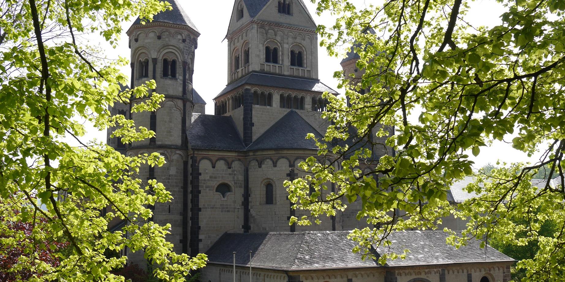 Durch die Bäume mit grünen Blättern ist die Abteikirche Maria Laach zu sehen.